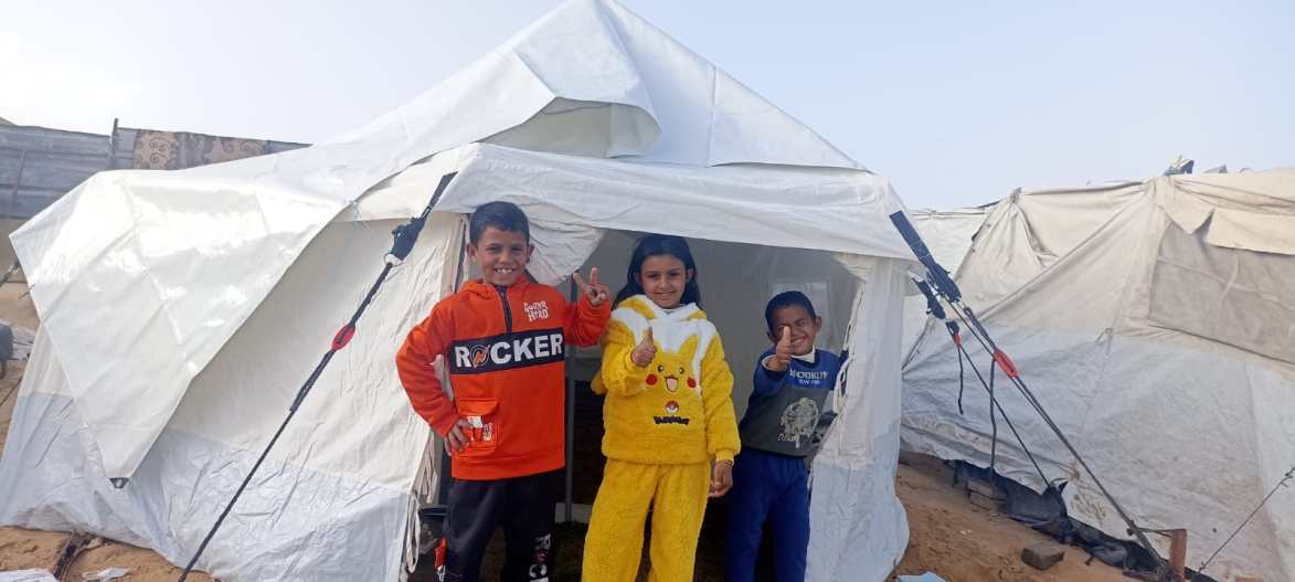 Children in warm clothing standing in front of a shelter tent in Gaza during winter
