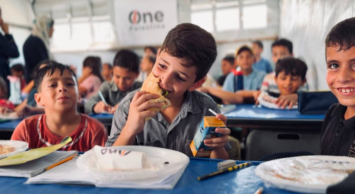 Children in a Gaza tent classroom smiling with One Palestine pizzas on their desks