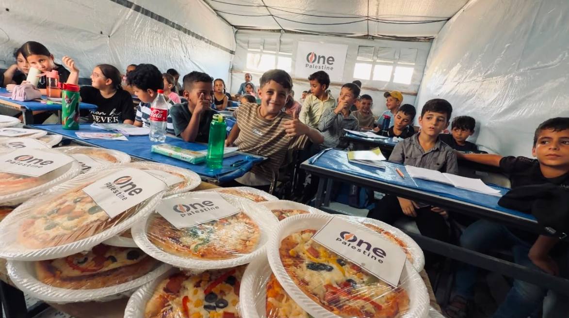 A boy in a Gaza tent classroom gives a thumbs up beside One Palestine branded pizzas